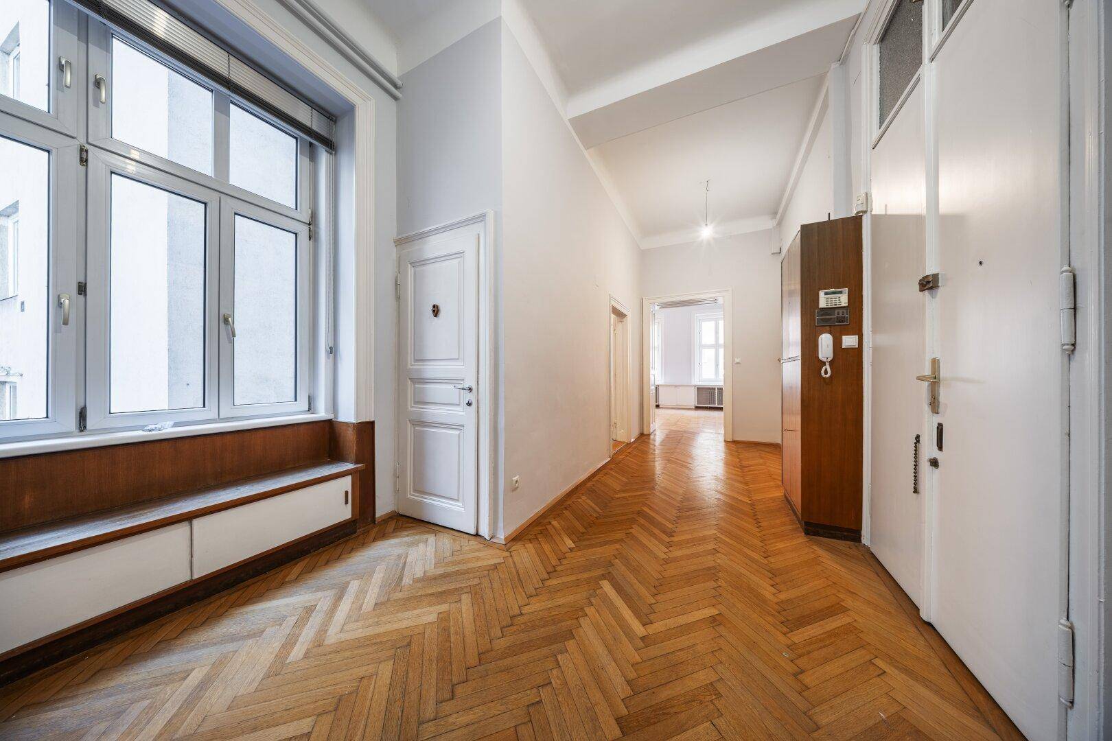 Langer Flur mit Fischgrätparkett, hohen Decken und Einbauschränken unter dem Fenster.