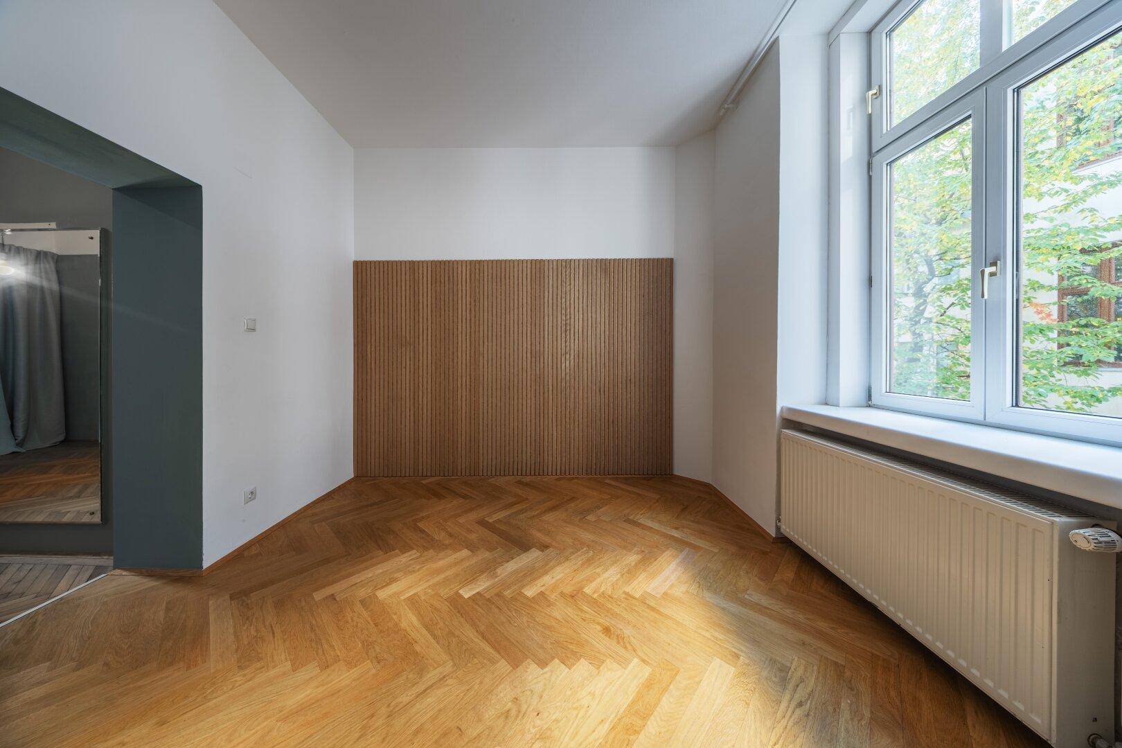 Geräumiges Zimmer mit Fischgrätparkett, Holzwandpaneelen und viel Tageslicht.