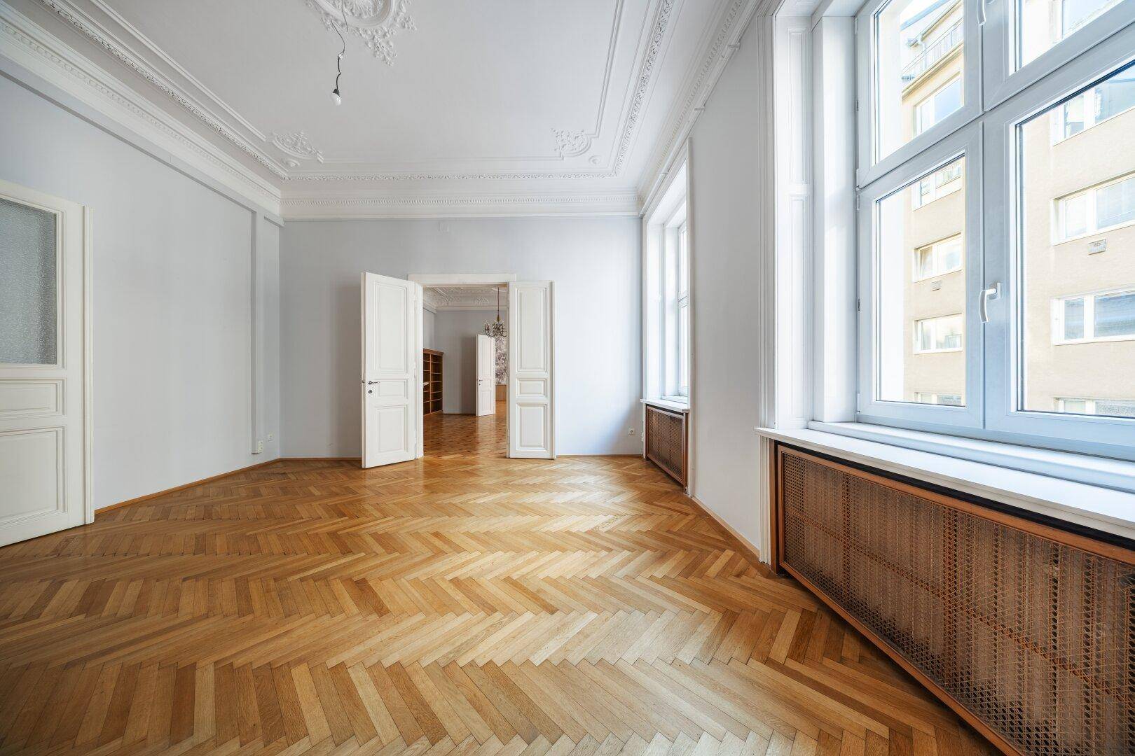 Heller Wohnraum mit Fischgrätparkett, hohen Decken und großen Fenstern mit Blick nach draußen.