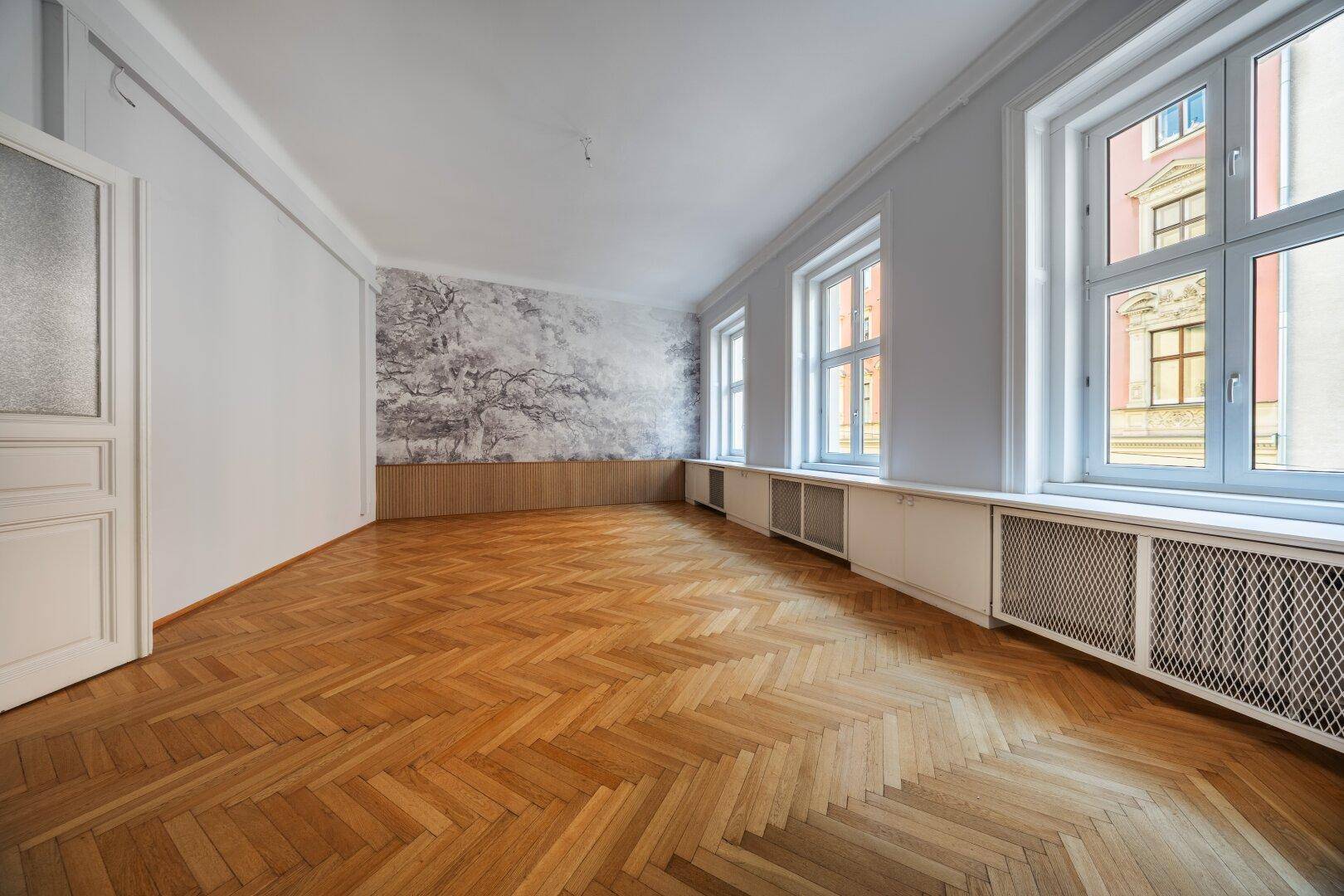 Helles Zimmer mit Fischgrätparkett, dekorativer Wandgestaltung und großen Fenstern.