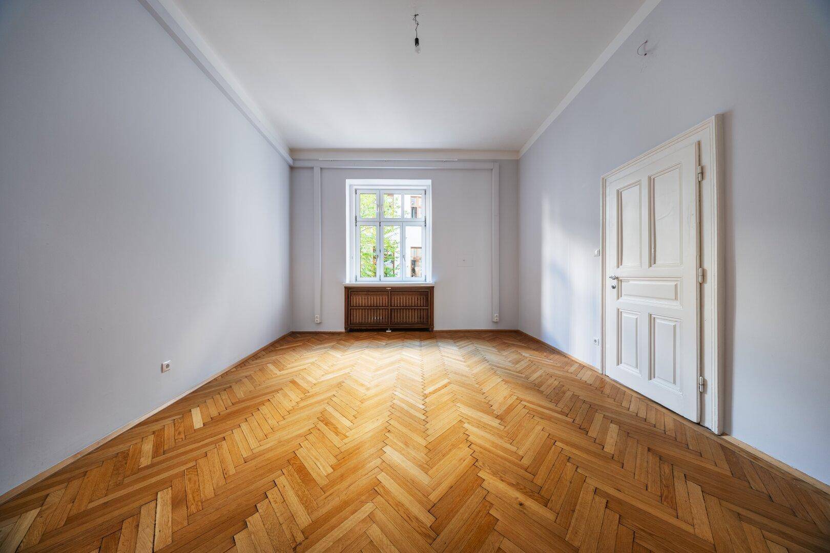 Ruhiges Zimmer mit Fischgrätparkett, hohen Decken und einem Fenster mit Blick ins Grüne.