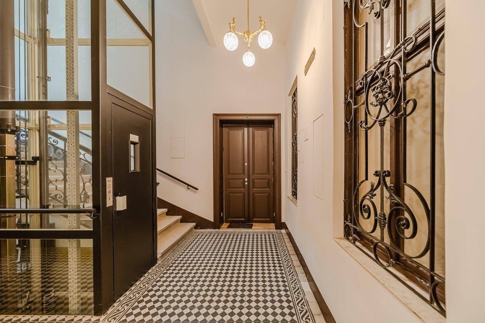 Eingangsbereich mit modernem Glasaufzug, Treppe und Schachbrettmuster-Fliesenboden.
