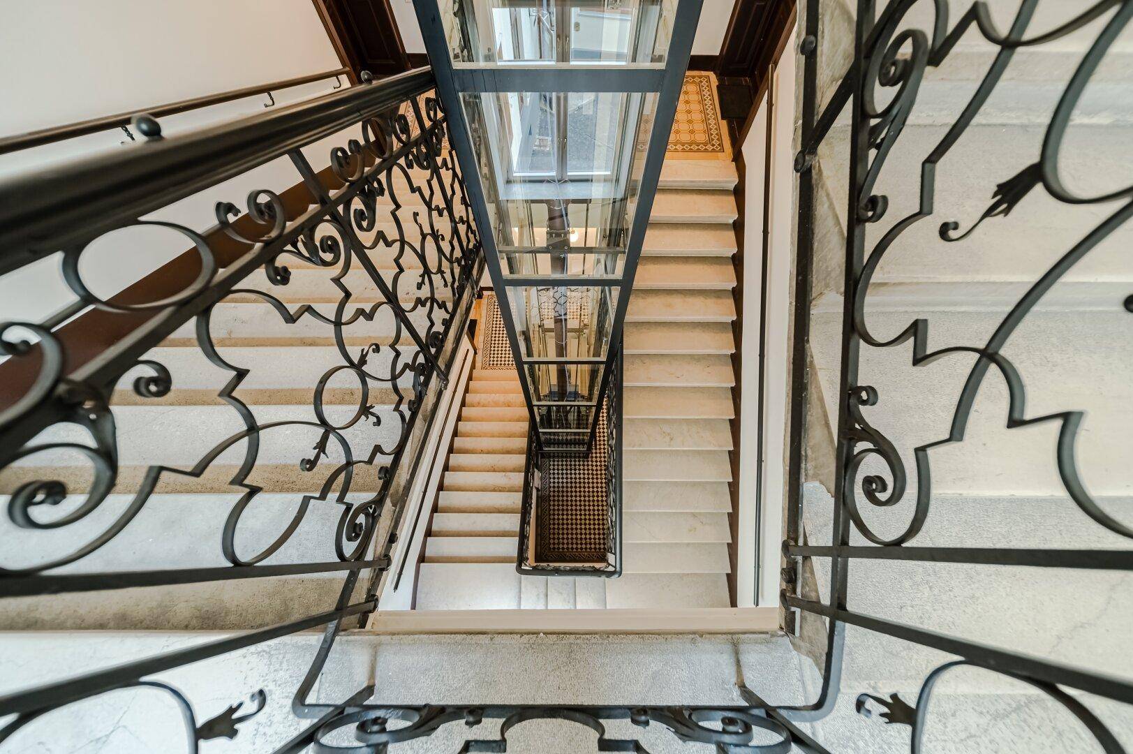 Elegantes Treppenhaus mit schmiedeeisernem Geländer und modernem Glasaufzug.