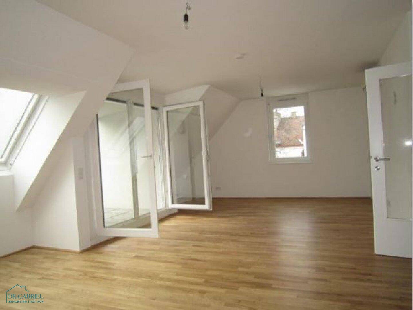Geräumiger, unmöblierter Raum mit Holzboden, Dachfenstern und Zugang zum Balkon.