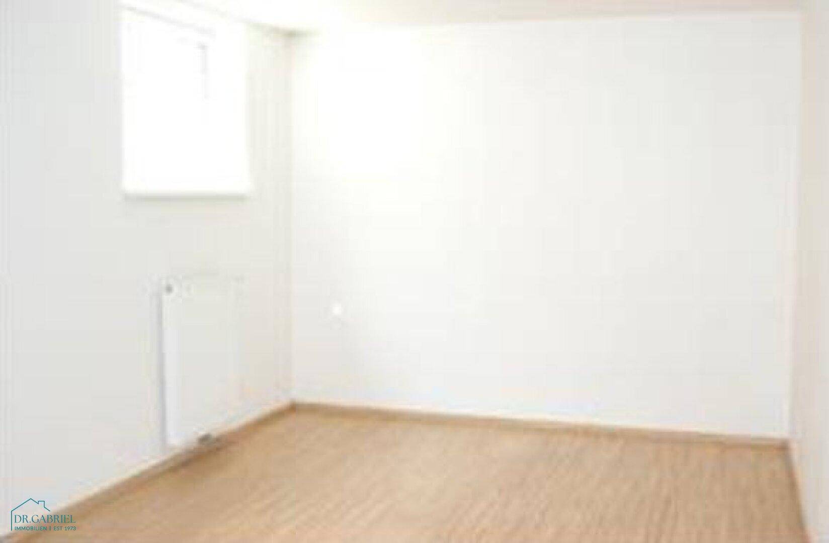 Einfaches, helles Zimmer mit Parkettboden und einem Fenster für Tageslicht.