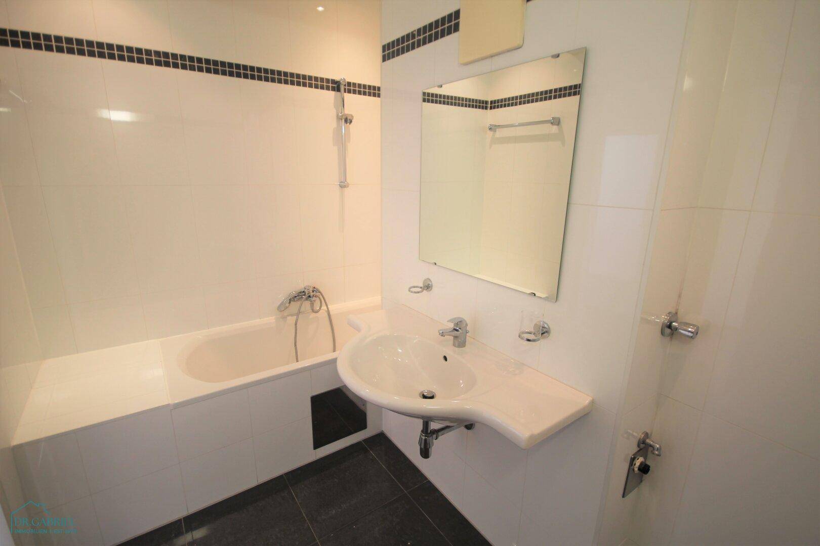 Helles Badezimmer mit Badewanne, Waschbecken und dunklen Bodenfliesen.