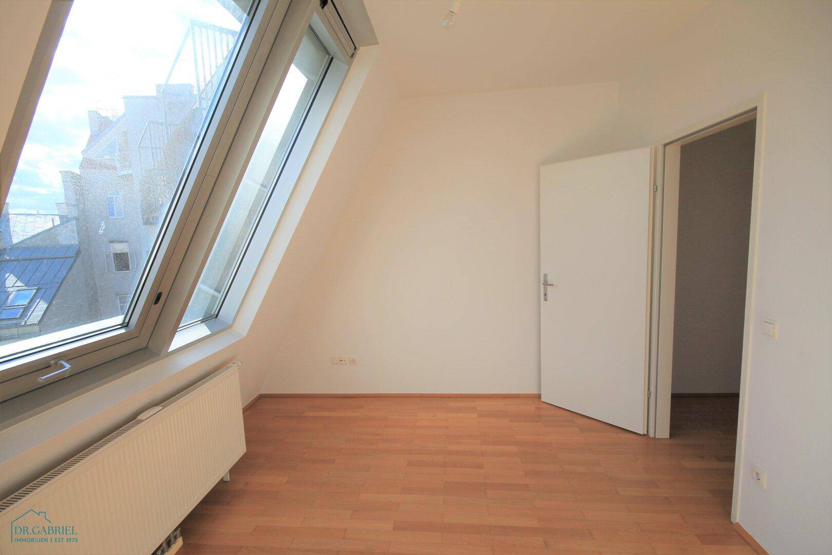 Helles Schlafzimmer mit Dachschräge, Parkettboden und Blick nach draußen.