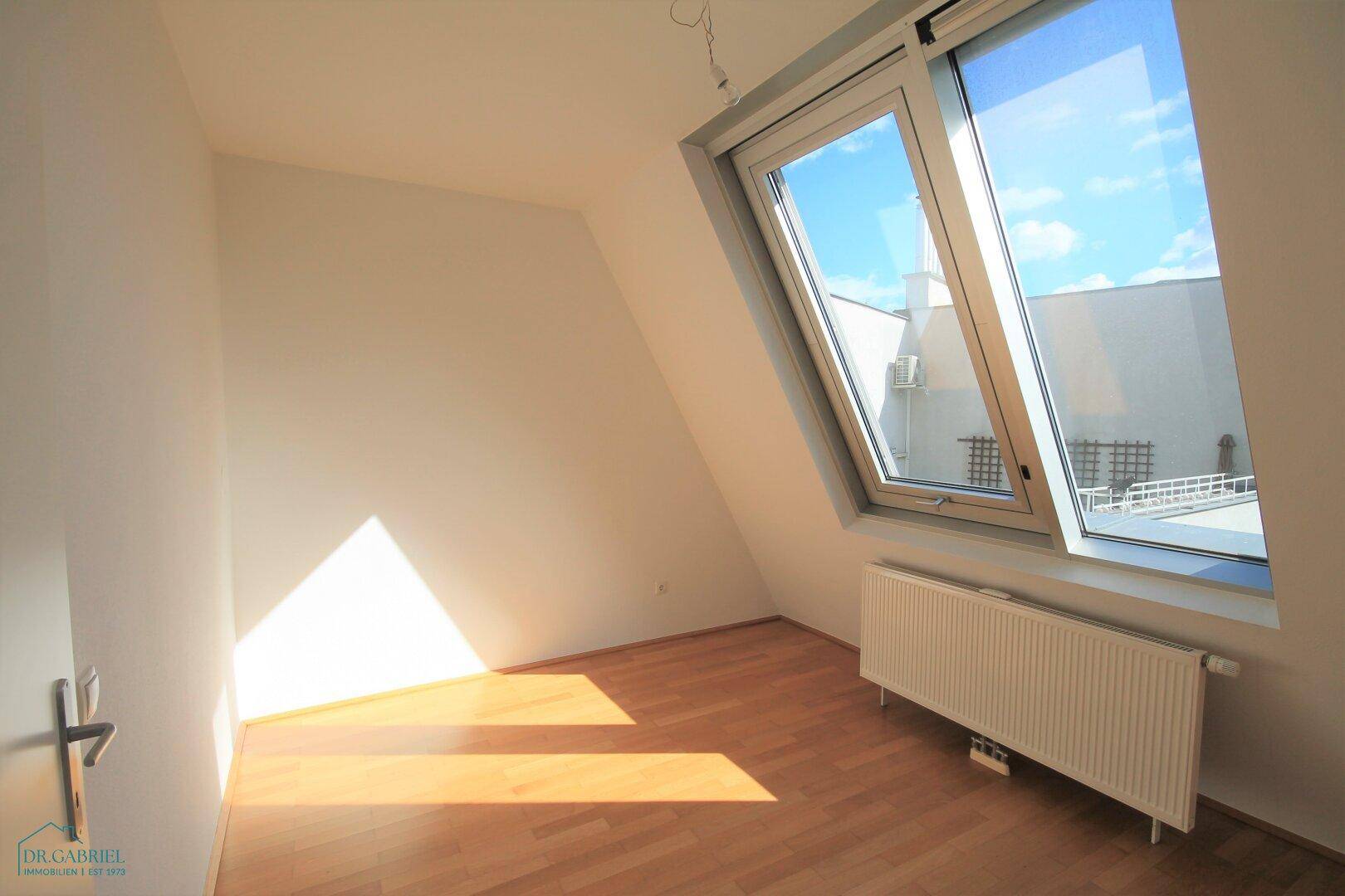 Sonniger Raum mit Dachschräge, Parkettboden und großem Fenster.