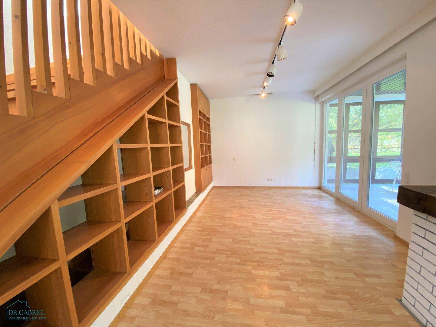 Heller Wohnbereich mit Holzboden, eingebautem Regal unter der Treppe und viel Tageslicht durch große Fenster.