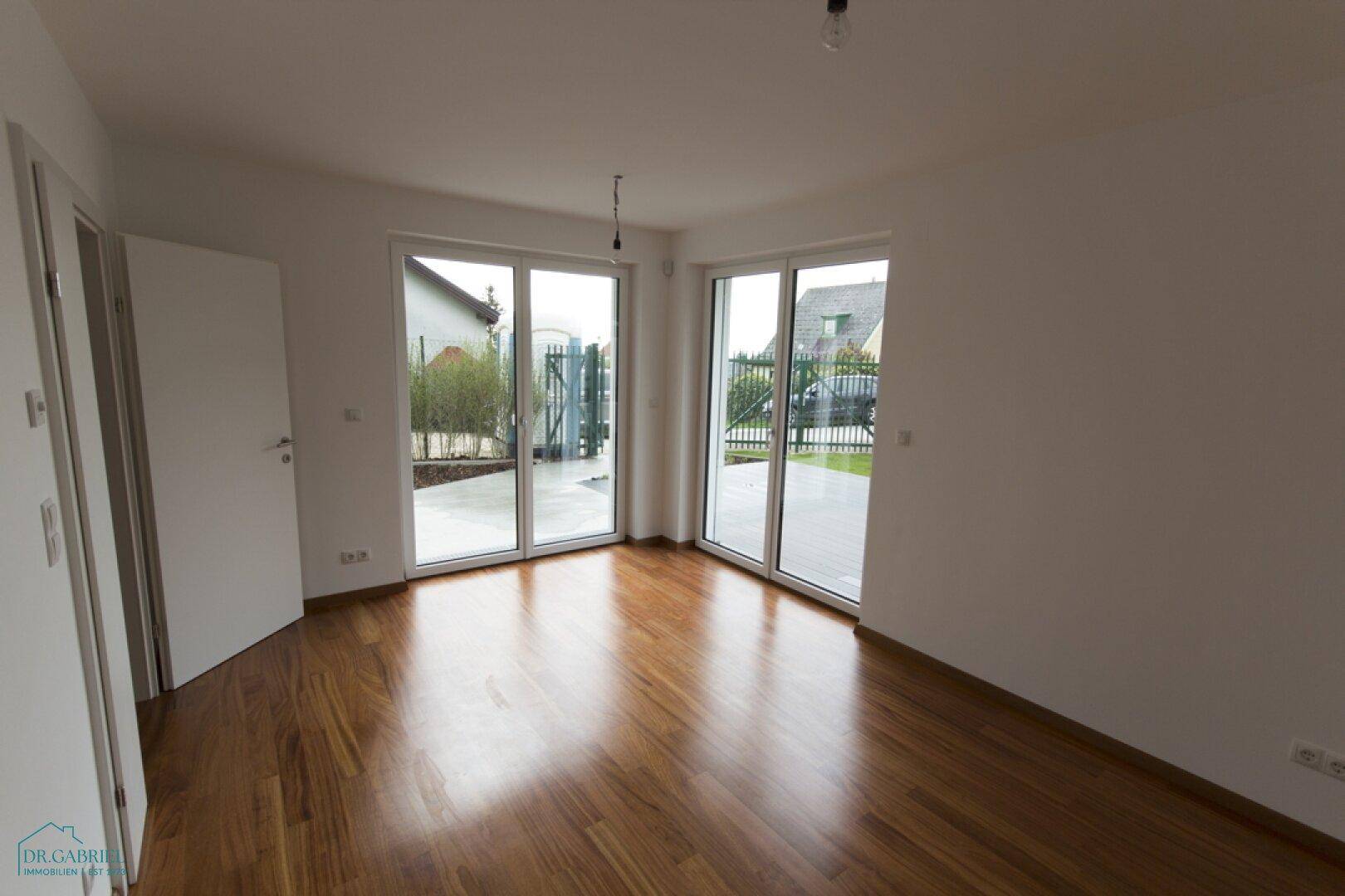 Heller Raum mit Holzboden und großen Glastüren, die Zugang zu einem gepflasterten Außenbereich bieten.