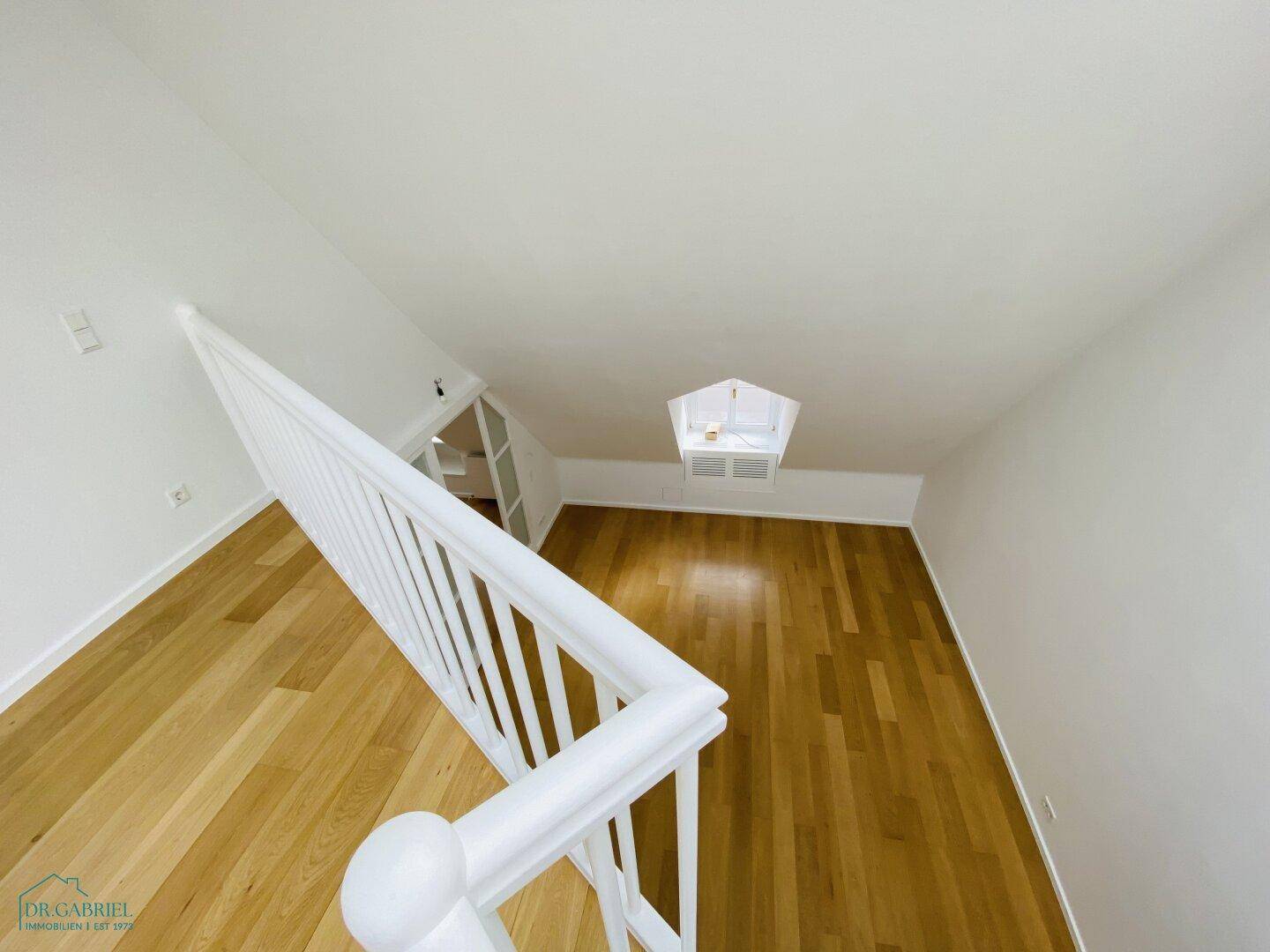 Blick von der Wendeltreppe in einen hellen Dachbodenraum mit Holzparkettboden und schrägen Wänden.