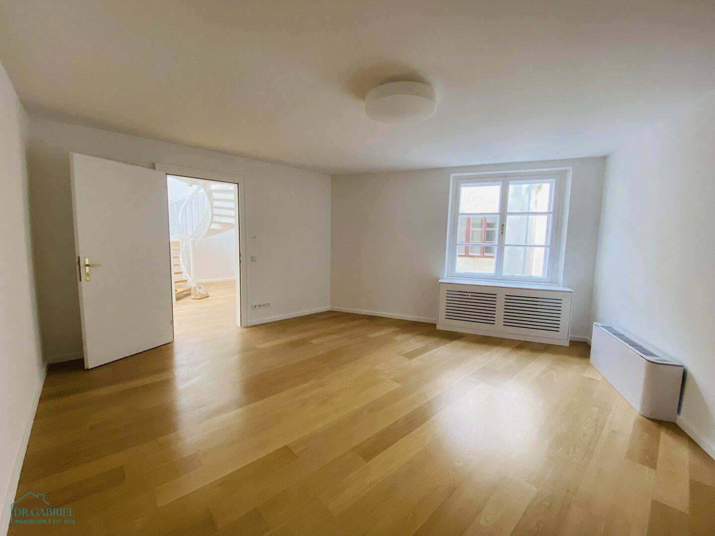 Helles Zimmer mit Holzparkettboden, weiß gestrichenen Wänden und einem Fenster mit Heizungsverkleidung.