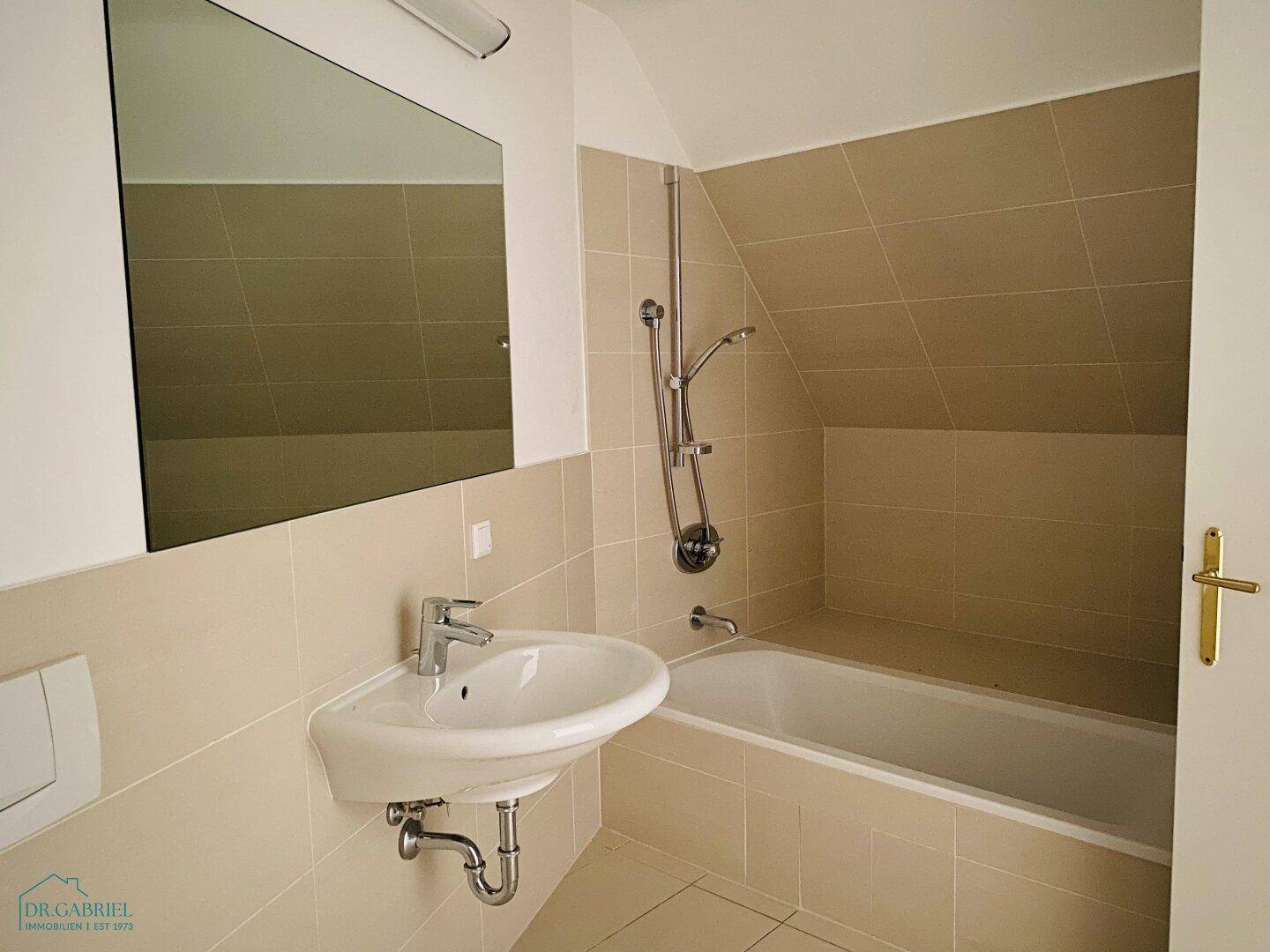 Modernes Badezimmer mit beigefarbenen Fliesen, Badewanne und Waschbecken unter einer Dachschräge.