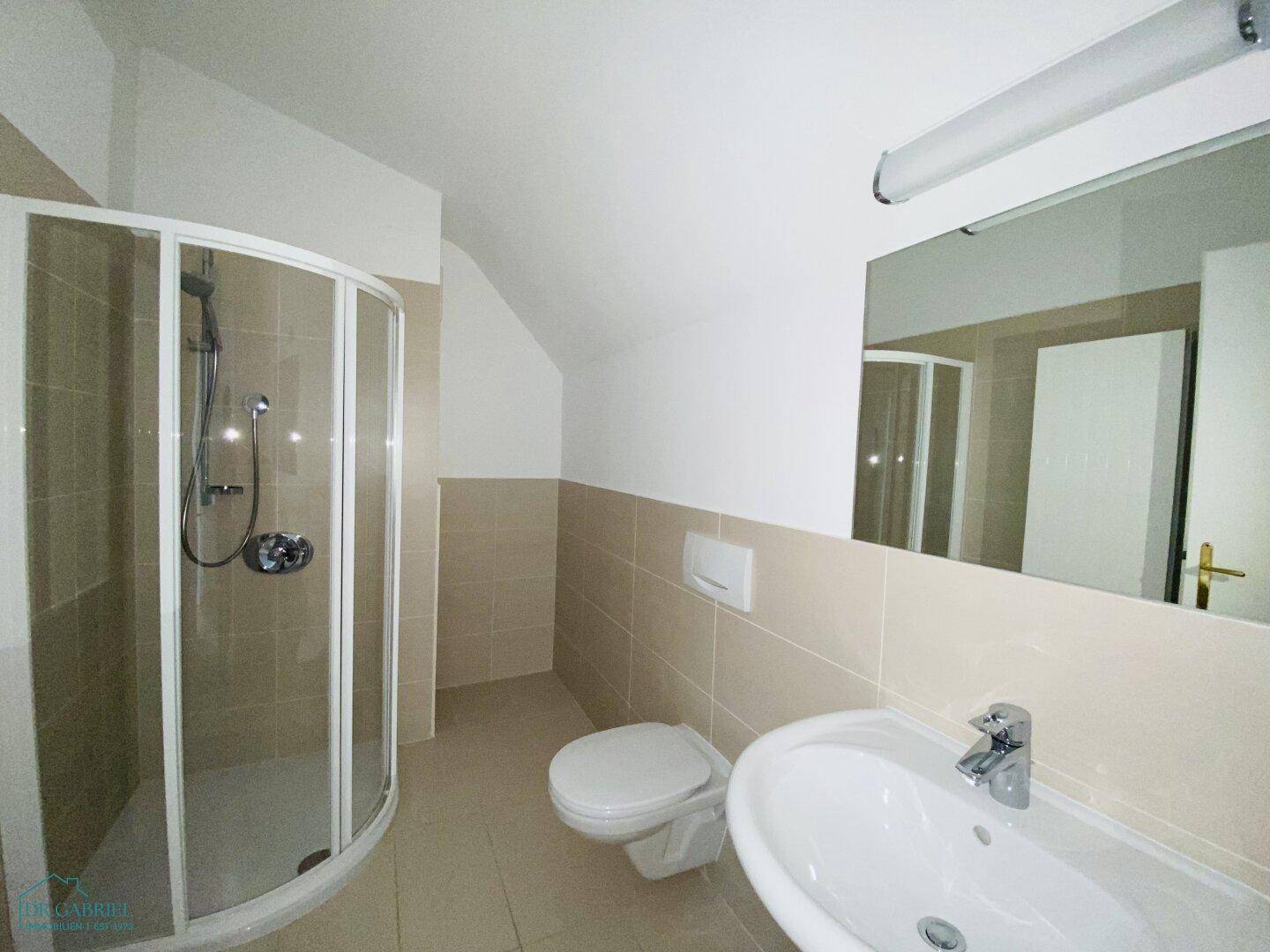 Modernes Badezimmer mit beigefarbenen Fliesen, Duschkabine, WC und Waschbecken unter einer Dachschräge.