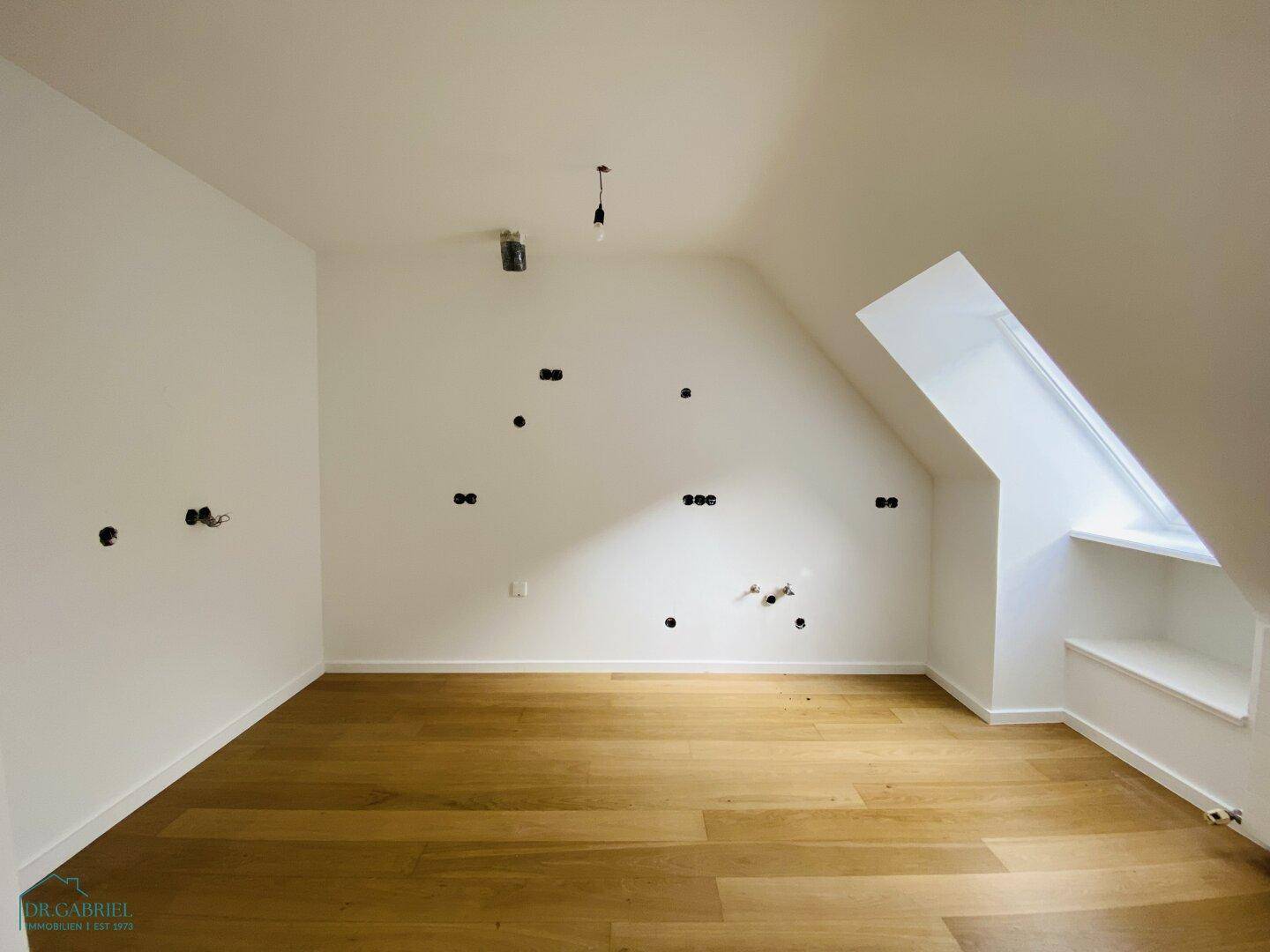 Unfertiger Dachbodenraum mit Holzparkettboden, schrägen Wänden und einem Dachfenster.