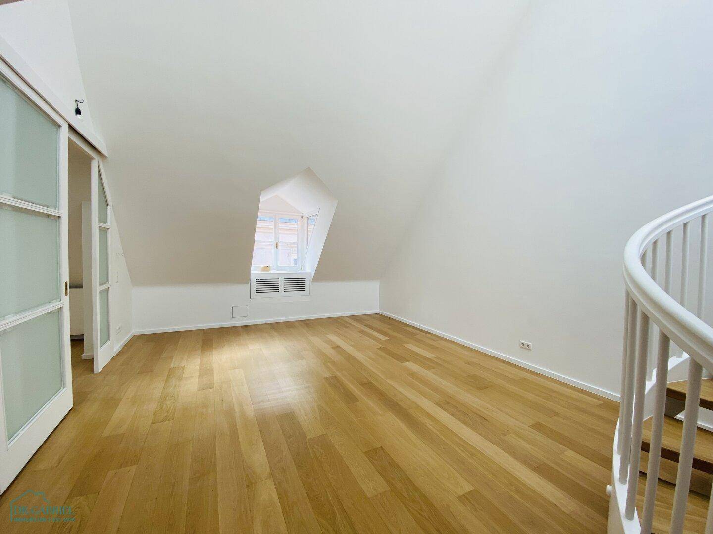 Heller Dachbodenraum mit Holzparkettboden, schrägen Wänden und einem kleinen Fenster.