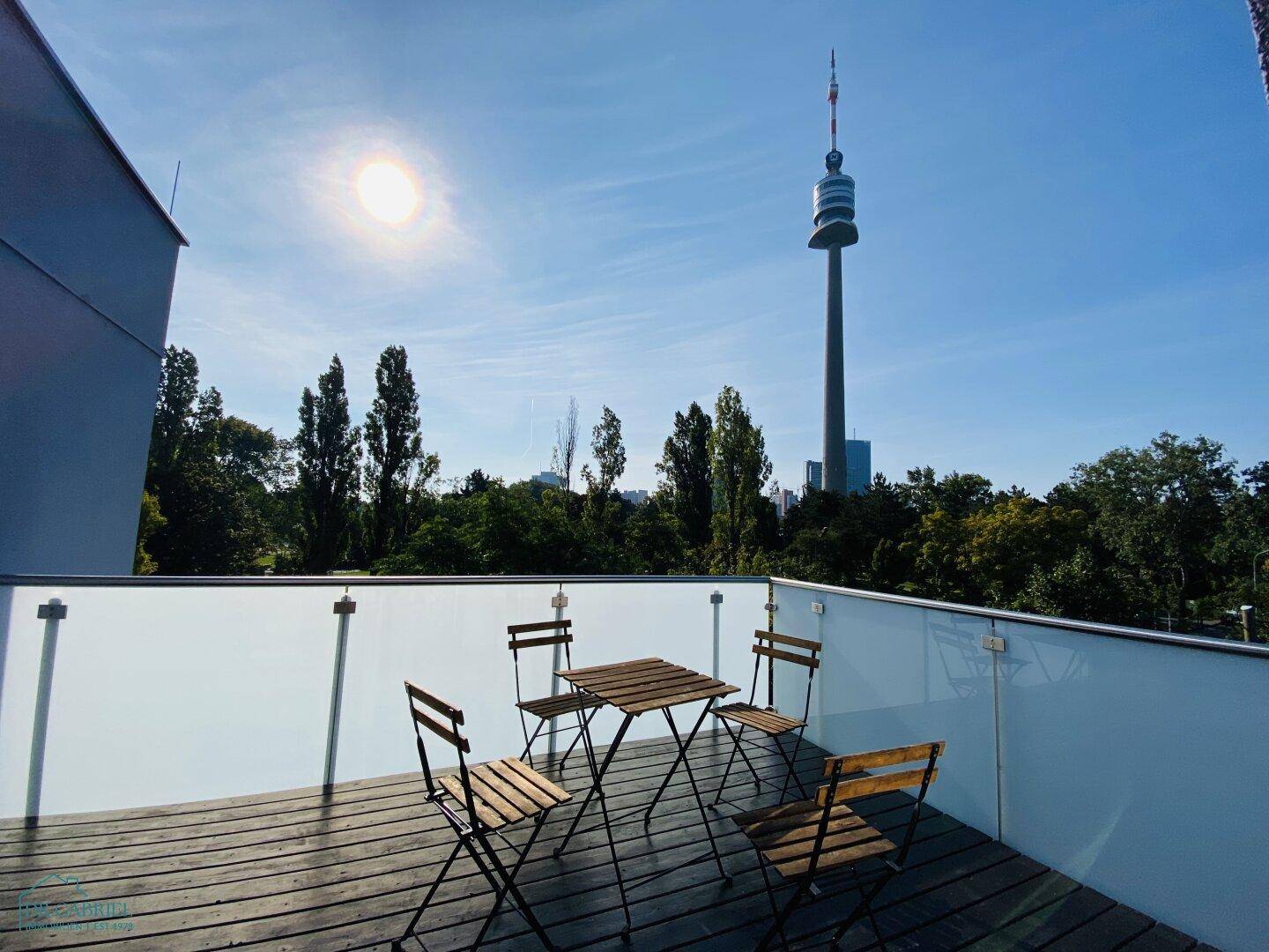 Sonnige Terrasse mit Holzboden und Sitzgelegenheiten, bietet einen weiten Blick auf den Donauturm.