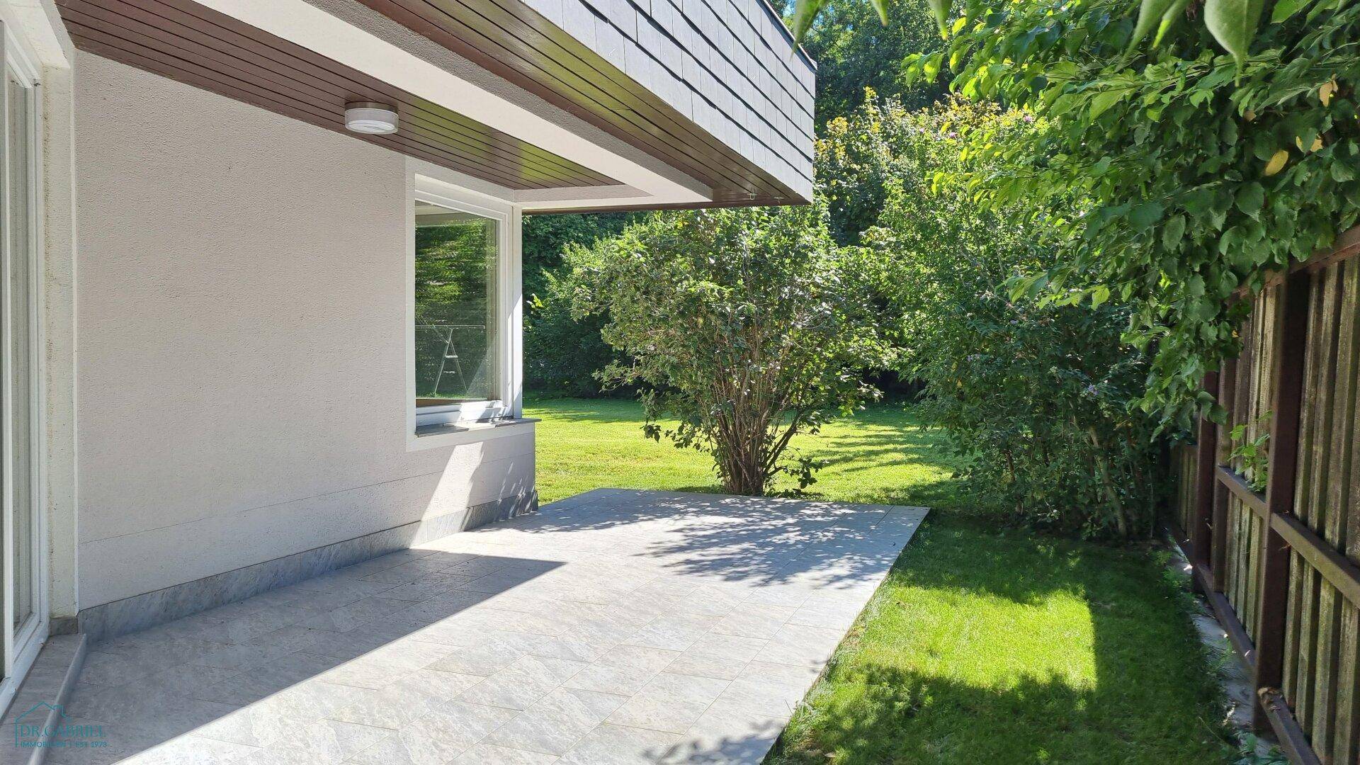 Geräumige, gepflasterte Terrasse am Haus, umgeben von Rasen und Sträuchern, ideal für den Außenbereich.