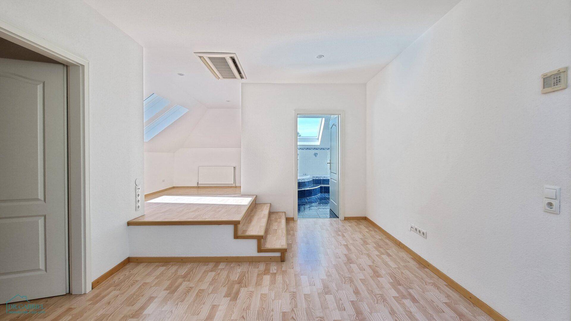 Heller Raum mit Holzfußboden, erhöhter Plattform und Zugang zu einem Badezimmer im Dachgeschoss.
