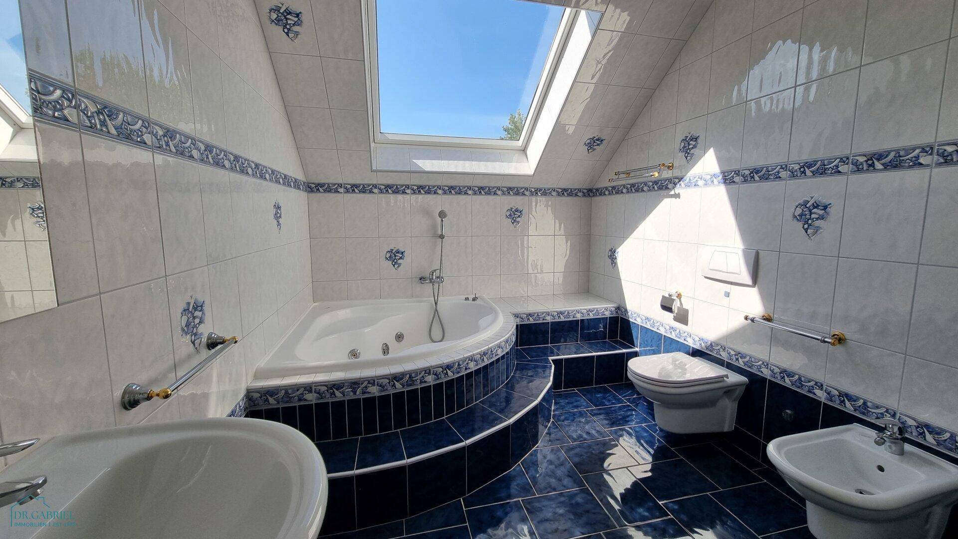 Geräumiges Badezimmer im Dachgeschoss mit blauen und weißen Fliesen, Eck-Whirlpool und großem Dachfenster.