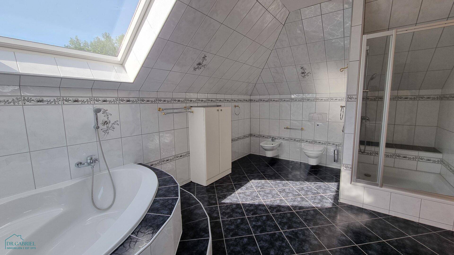 Geräumiges Badezimmer mit dunklen Bodenfliesen, Eckbadewanne, Dusche und Dachfenster für natürliches Licht.