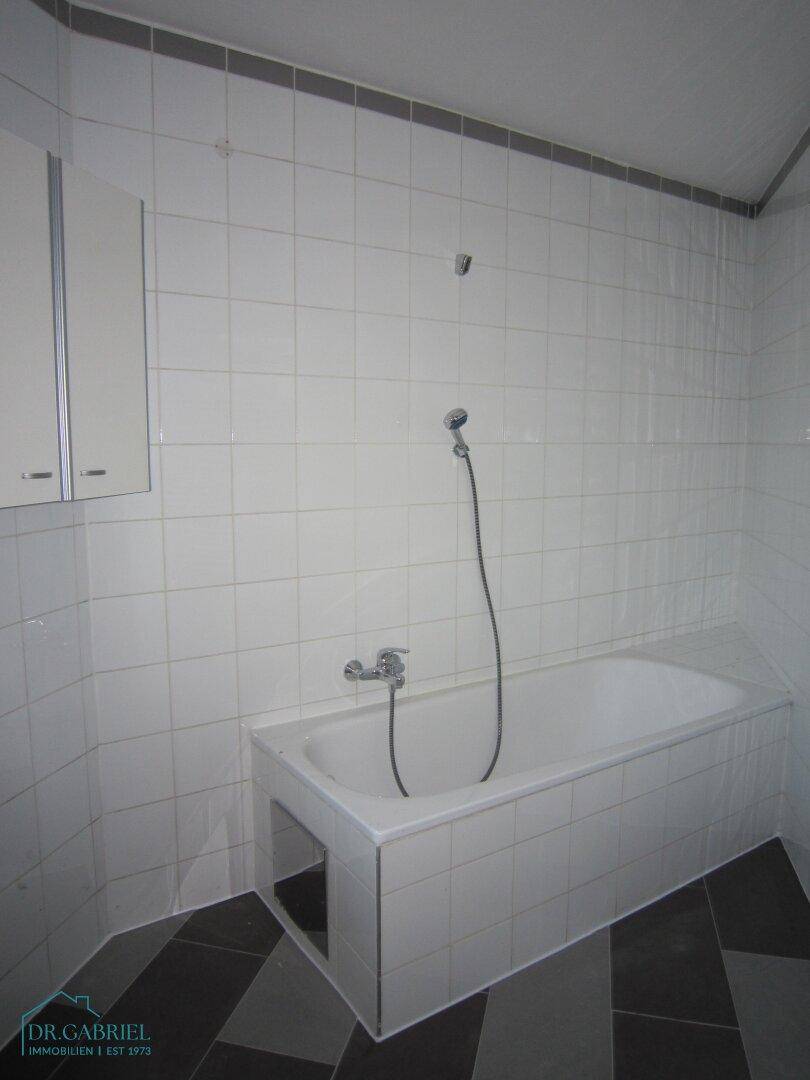 Badezimmer mit weißen Wandfliesen, gemustertem Boden und einer Badewanne mit Duschfunktion.