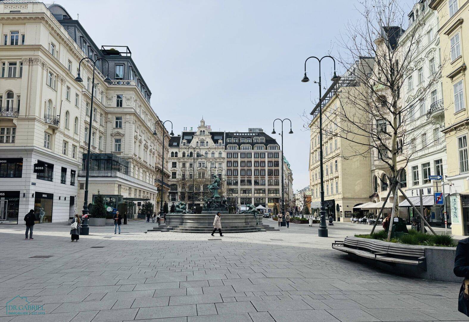 Weitläufiger Stadtplatz mit historischen Gebäuden, einem Brunnen in der Mitte und Menschen, die spazieren gehen.