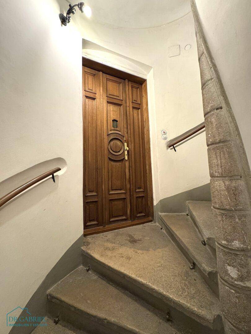 Historisches Treppenhaus mit Steinstufen, Holzgeländer und einer verzierten Holztür am oberen Ende.