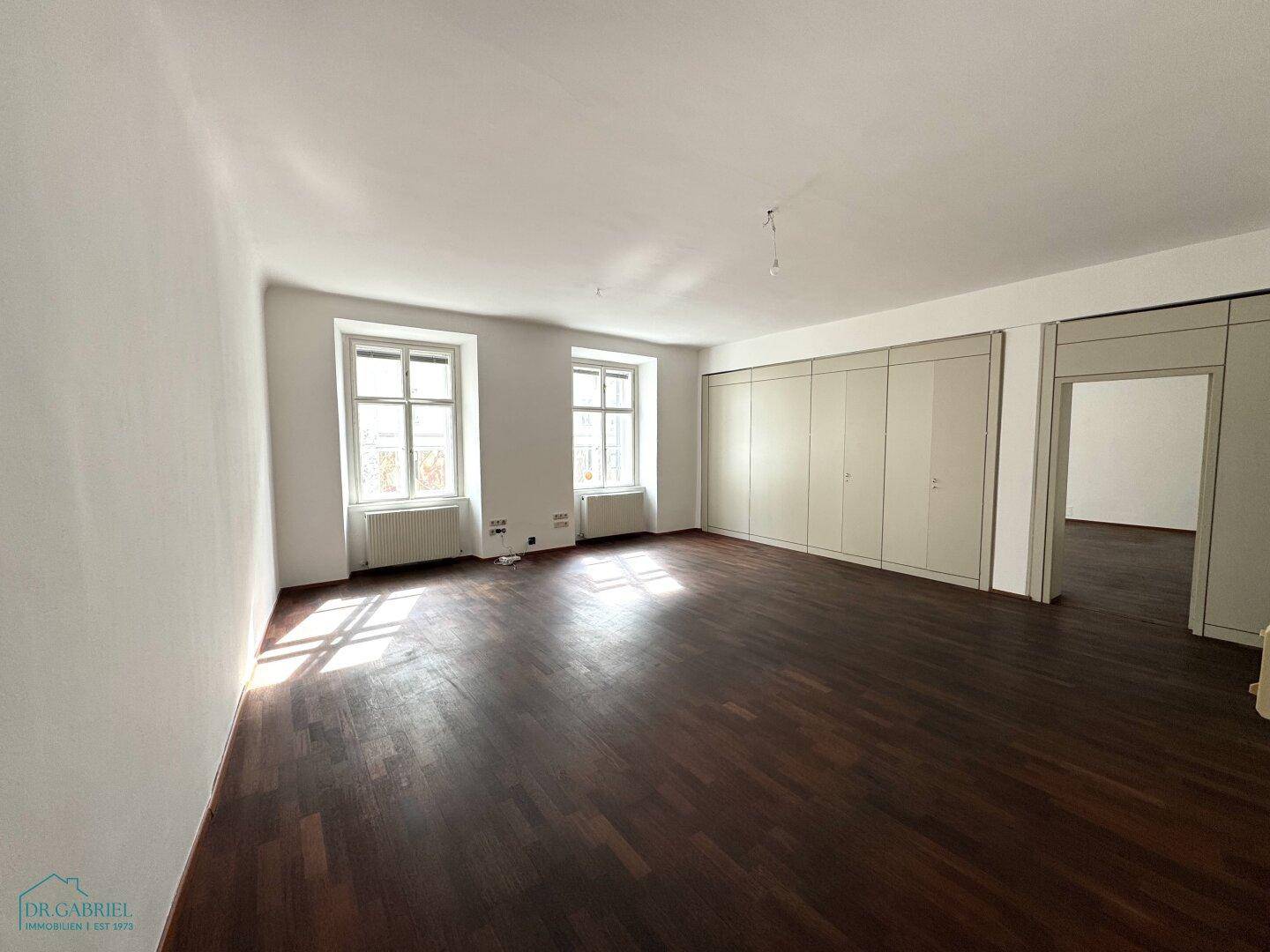 Helles, leeres Büro mit dunklem Holzboden, weißen Wänden und zwei großen Fenstern, die den Raum erhellen.