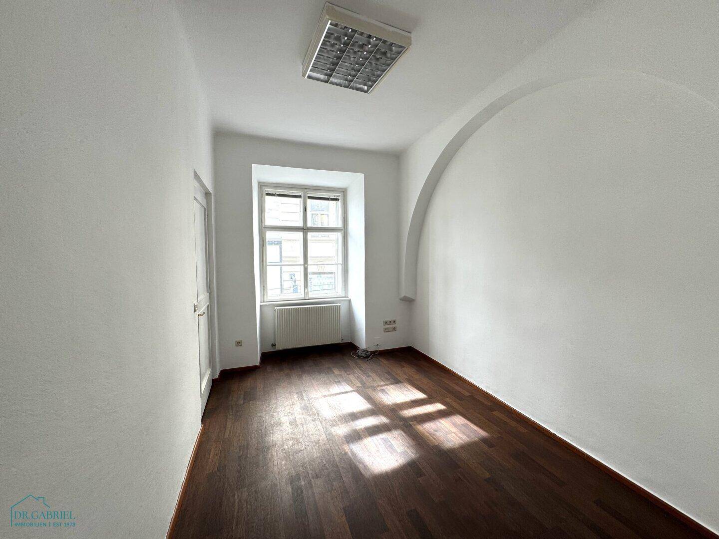 Kleineres, leeres Zimmer mit dunklem Holzboden, weißer Wand und einem Fenster, das Tageslicht spendet.