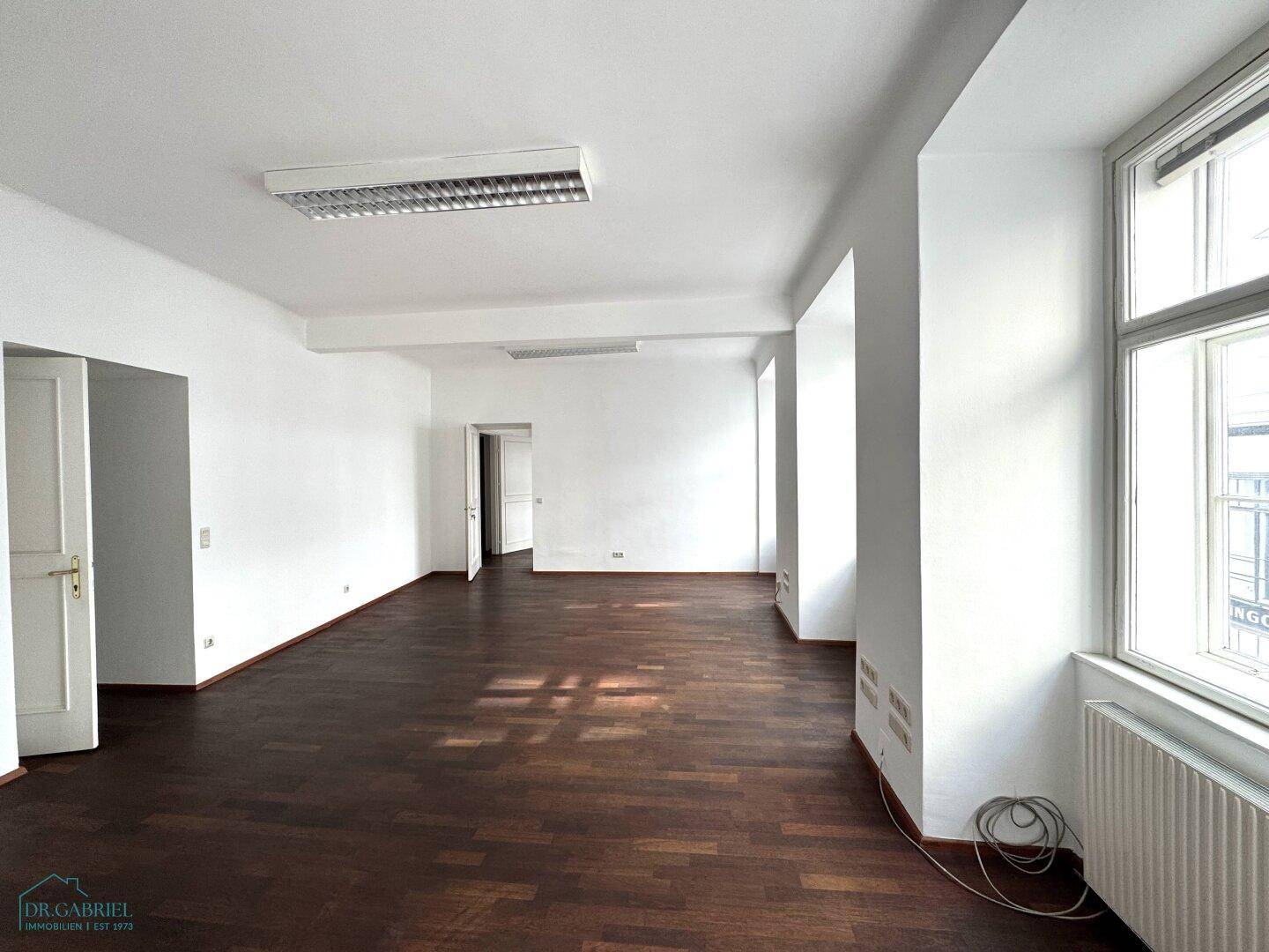 Geräumiges, leeres Büro mit dunklem Holzboden, weißen Wänden und hohen Fenstern, die viel Licht spenden.