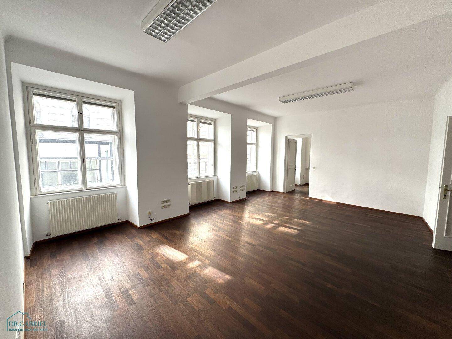Helles, leeres Büro mit dunklem Holzboden, weißen Wänden und mehreren Fenstern, die den Raum erhellen.