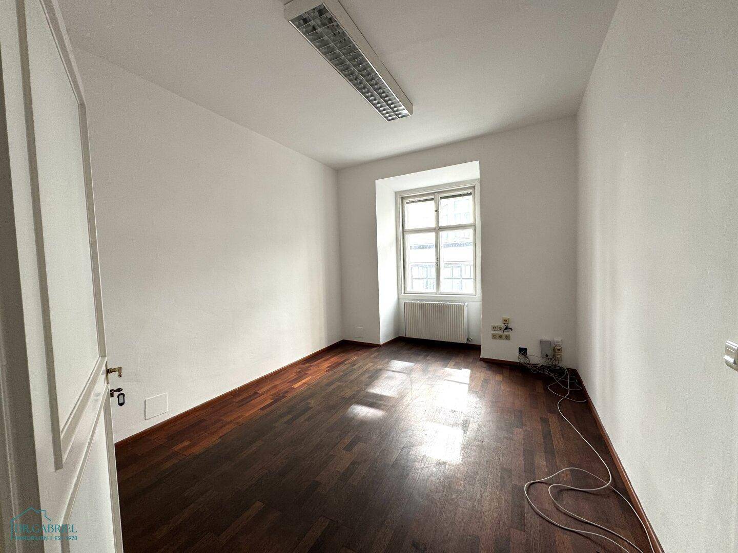 Heller Raum mit dunklem Holzfußboden, weißen Wänden und einem Fenster mit Heizkörper darunter.