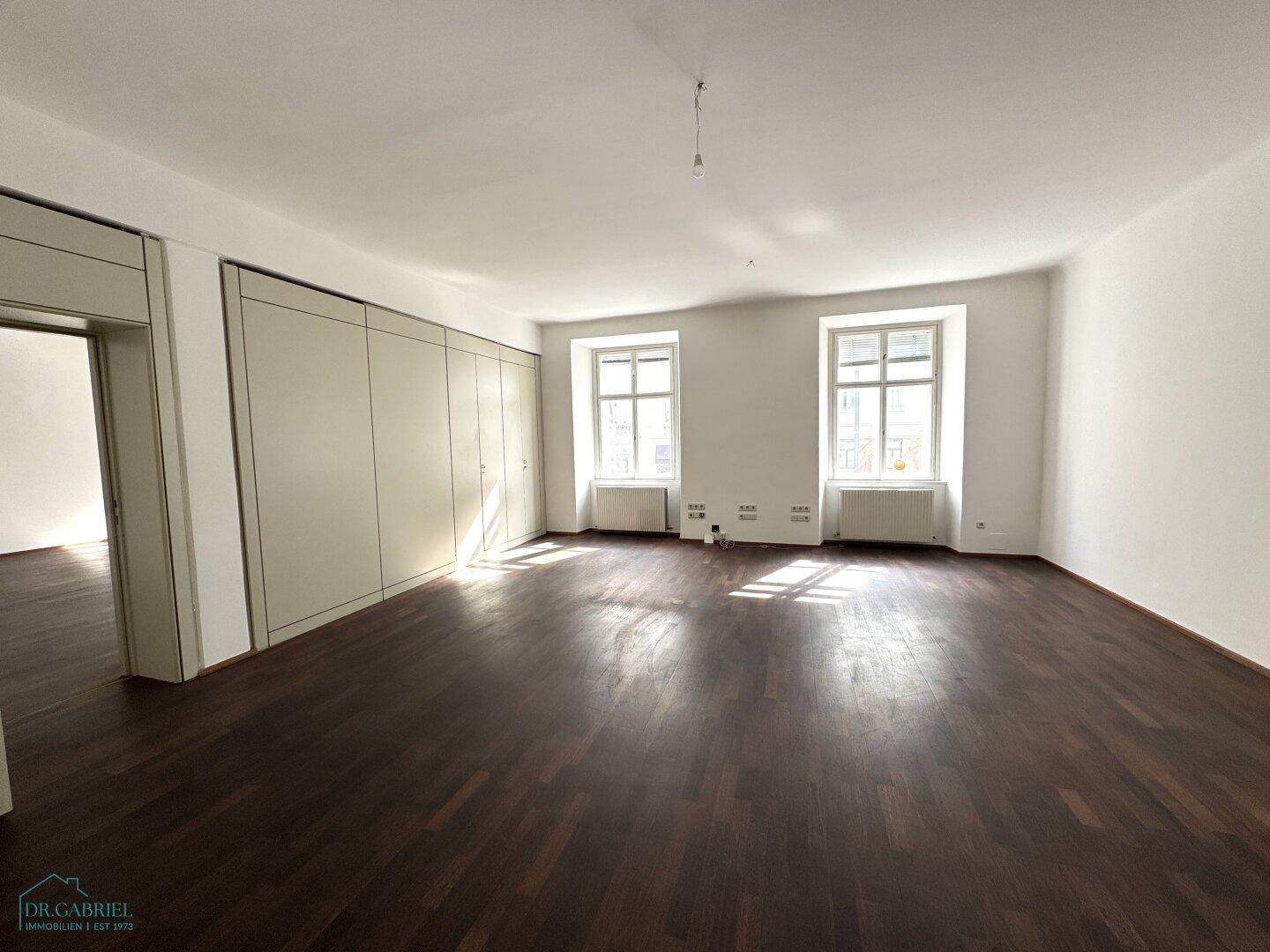Geräumiges, leeres Büro mit dunklem Holzboden, weißen Wänden und zwei großen Fenstern, die viel Tageslicht hereinlassen.
