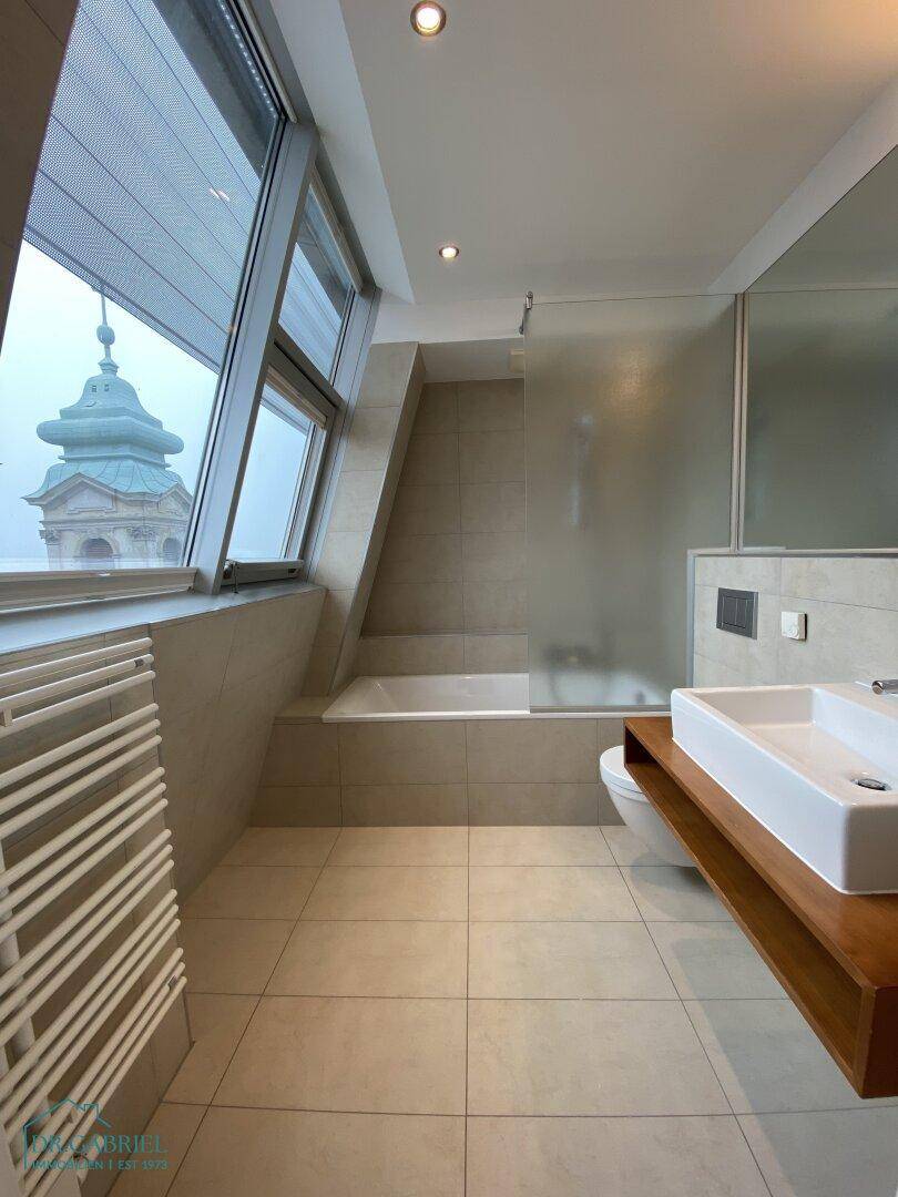 Modernes Badezimmer mit Badewanne, beigefarbenen Fliesen und schrägen Fenstern mit Blick auf Kirchtürme.