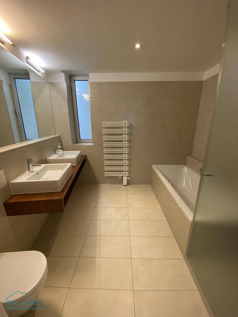 Modernes Badezimmer mit beigefarbenen Fliesen, Doppelwaschbecken, Badewanne und beheiztem Handtuchhalter.