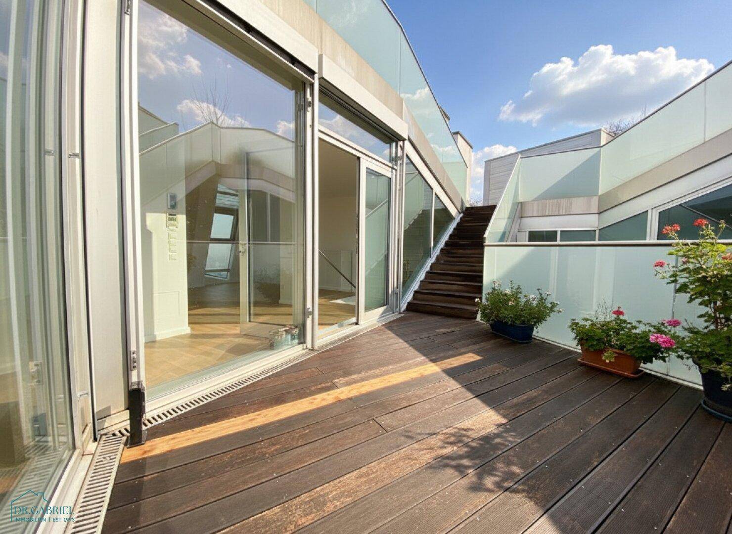 Sonnige Terrasse mit Holzboden und großen Glasfronten, die den Innenraum mit dem Außenbereich verbinden.