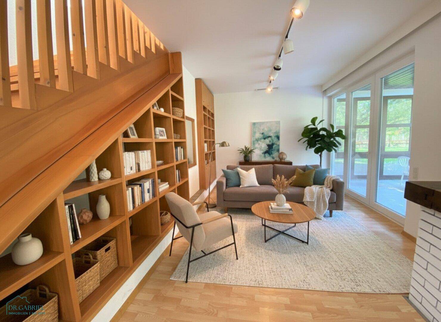 Gemütliches Wohnzimmer mit Holzboden, integriertem Bücherregal unter der Treppe und großen Fenstern.