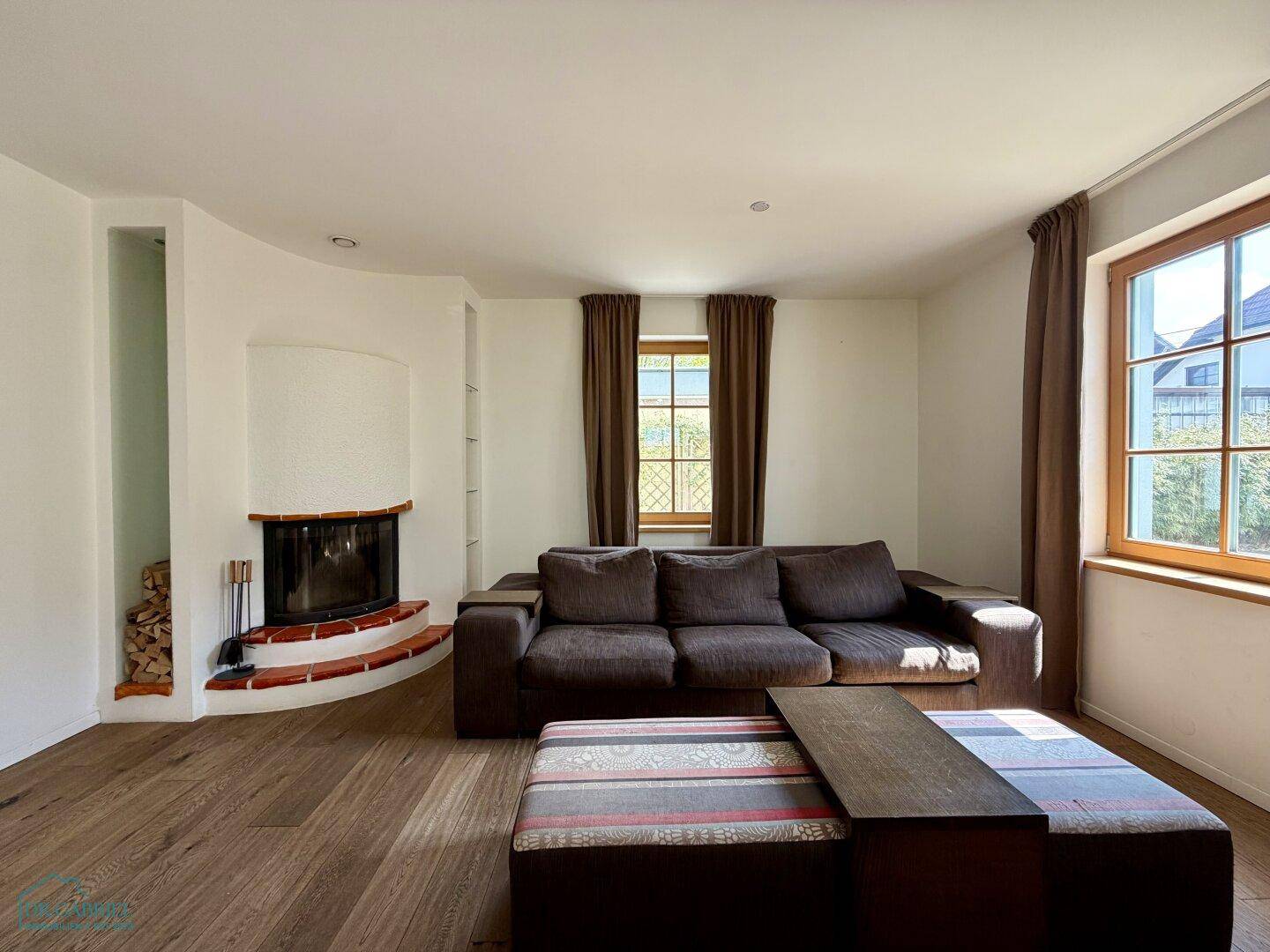 Gemütliches Wohnzimmer mit Kamin, Holzboden und großem Sofa, hell durch Fenster.