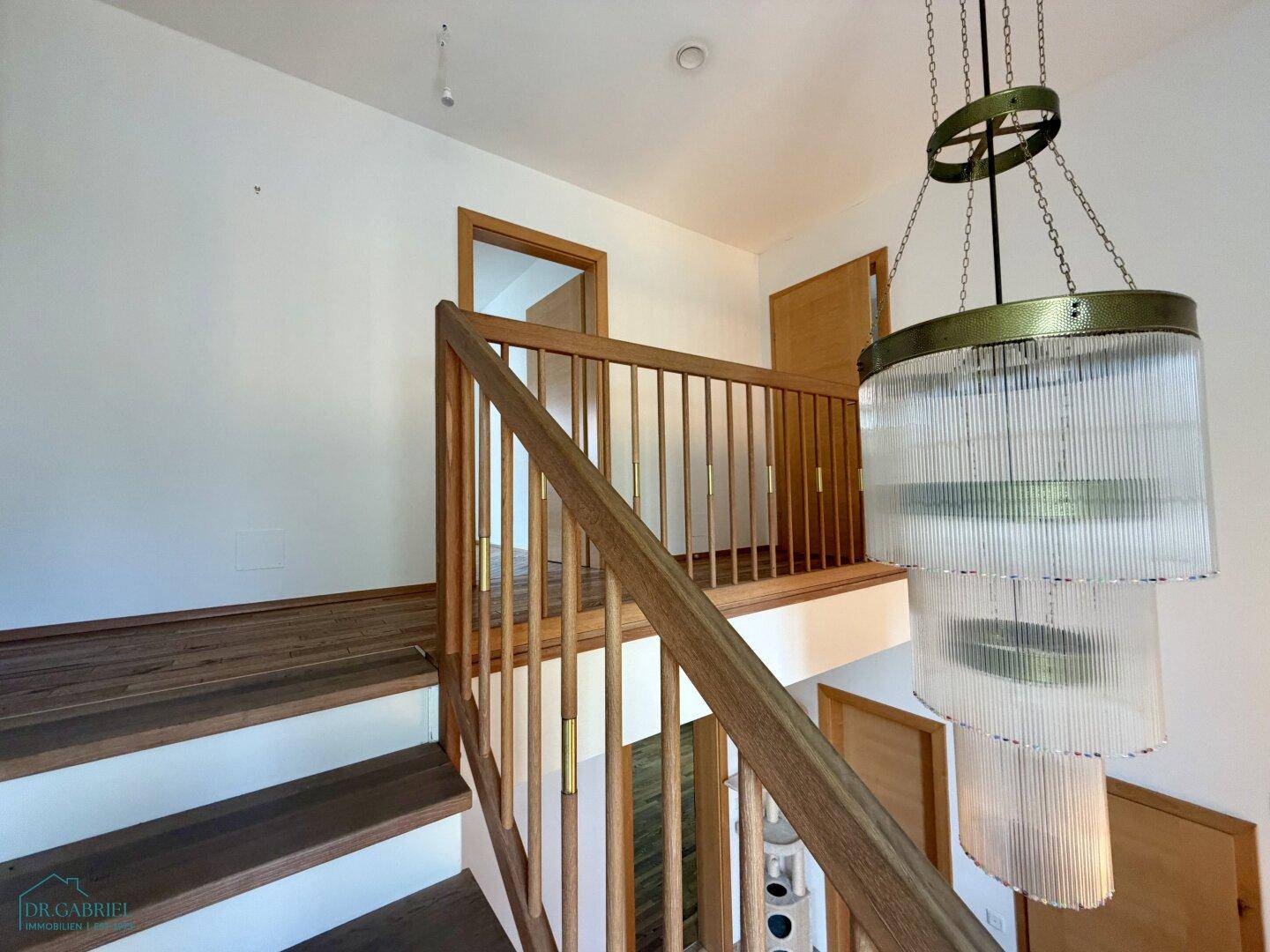 Treppenhaus mit Holzgeländer und moderner Hängelampe, führt zu den oberen Etagen.