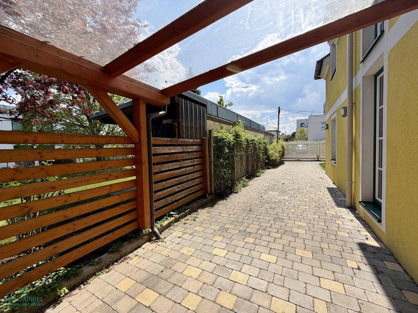 Gepflasterte Einfahrt mit Carport und Holzzaun, führt zum Haus.