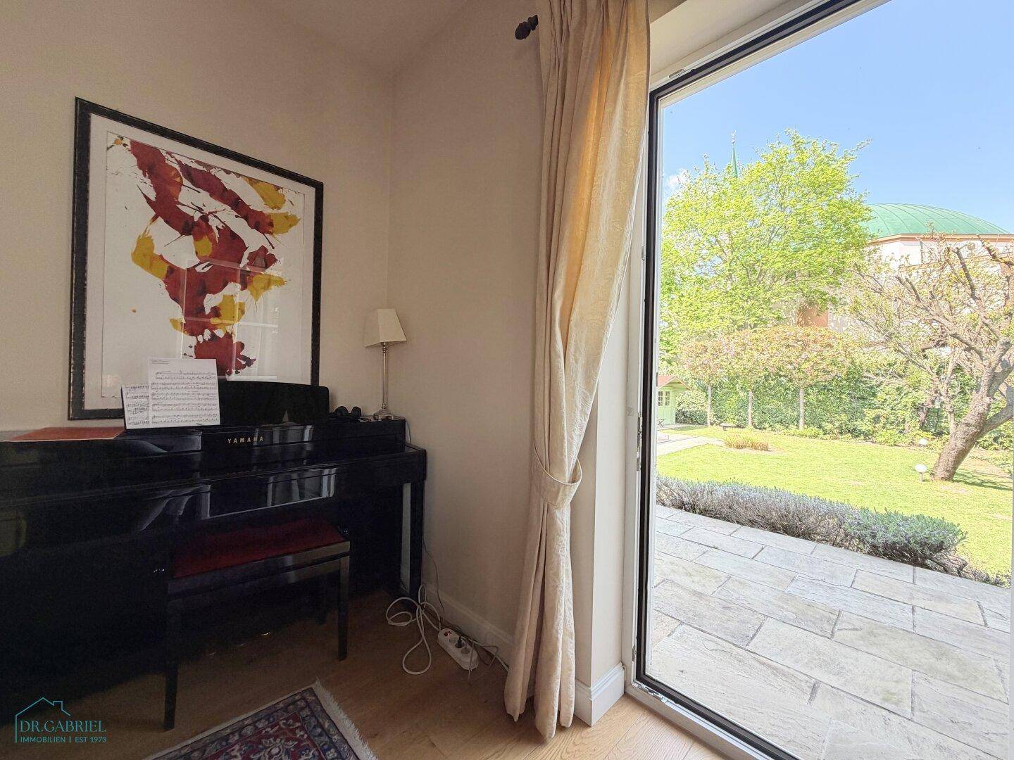 Heller Raum mit Klavier, Holzboden und großem Fenster mit Blick in den grünen Garten.