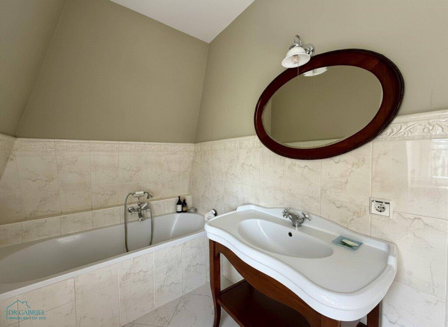 Klassisches Badezimmer mit Badewanne, elegantem Waschtisch und ovalem Spiegel.