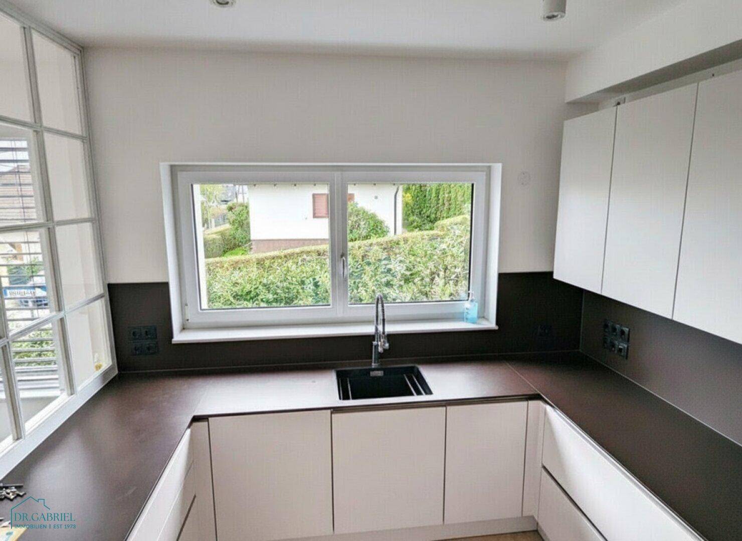Moderne Küche mit weißen Unterschränken, dunkler Arbeitsplatte und Fenster mit Blick ins Grüne.