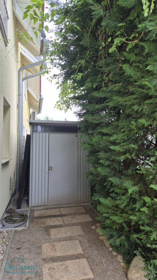 Seitenansicht des Hauses mit einem Gartenweg, umgeben von üppigem Grün und einem kleinen Schuppen.