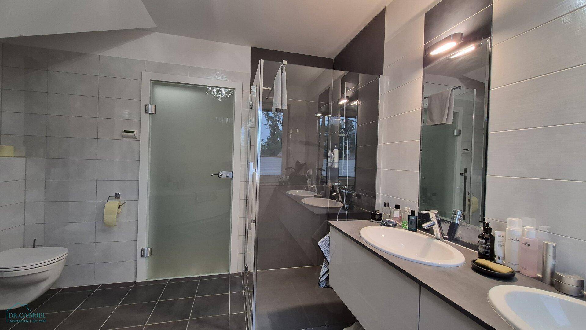 Modernes Badezimmer mit Dusche, Toilette und Doppelwaschbecken, in Grautönen gehalten.