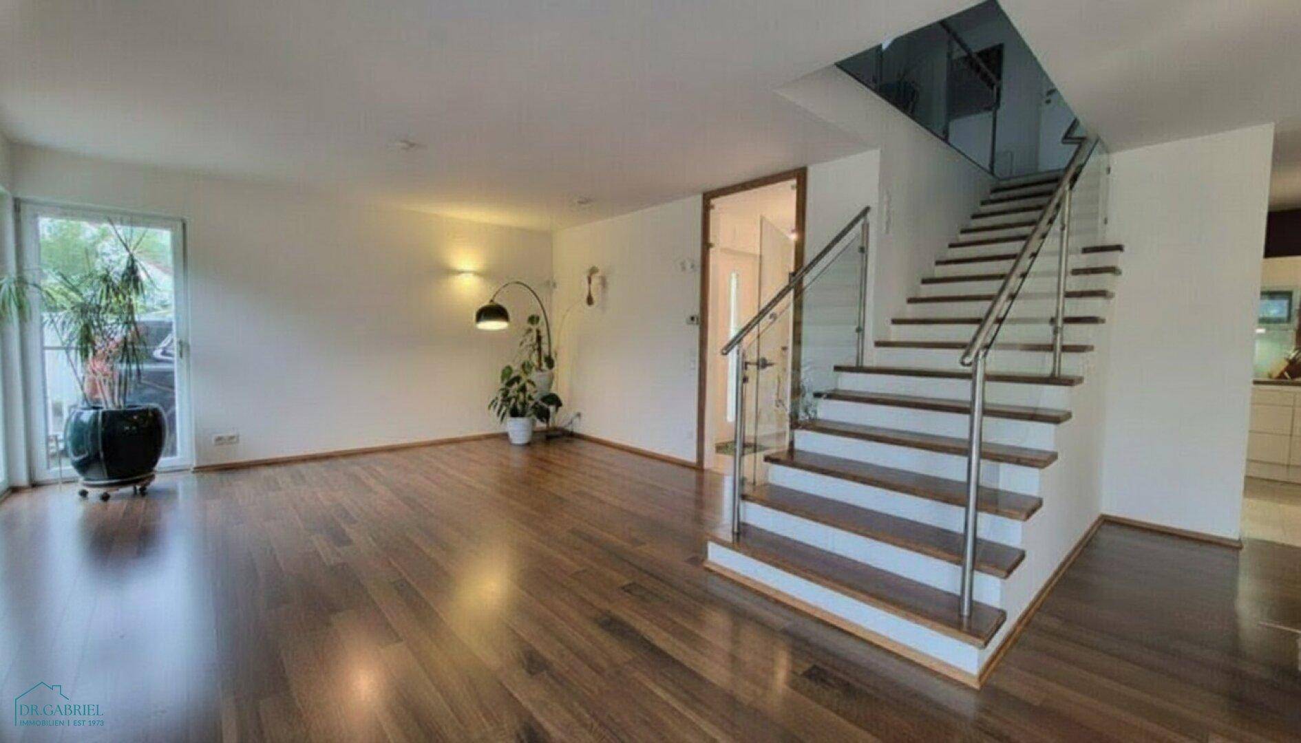 Geräumiges Wohnzimmer mit dunklem Holzboden und moderner Treppe mit Glasgeländer.