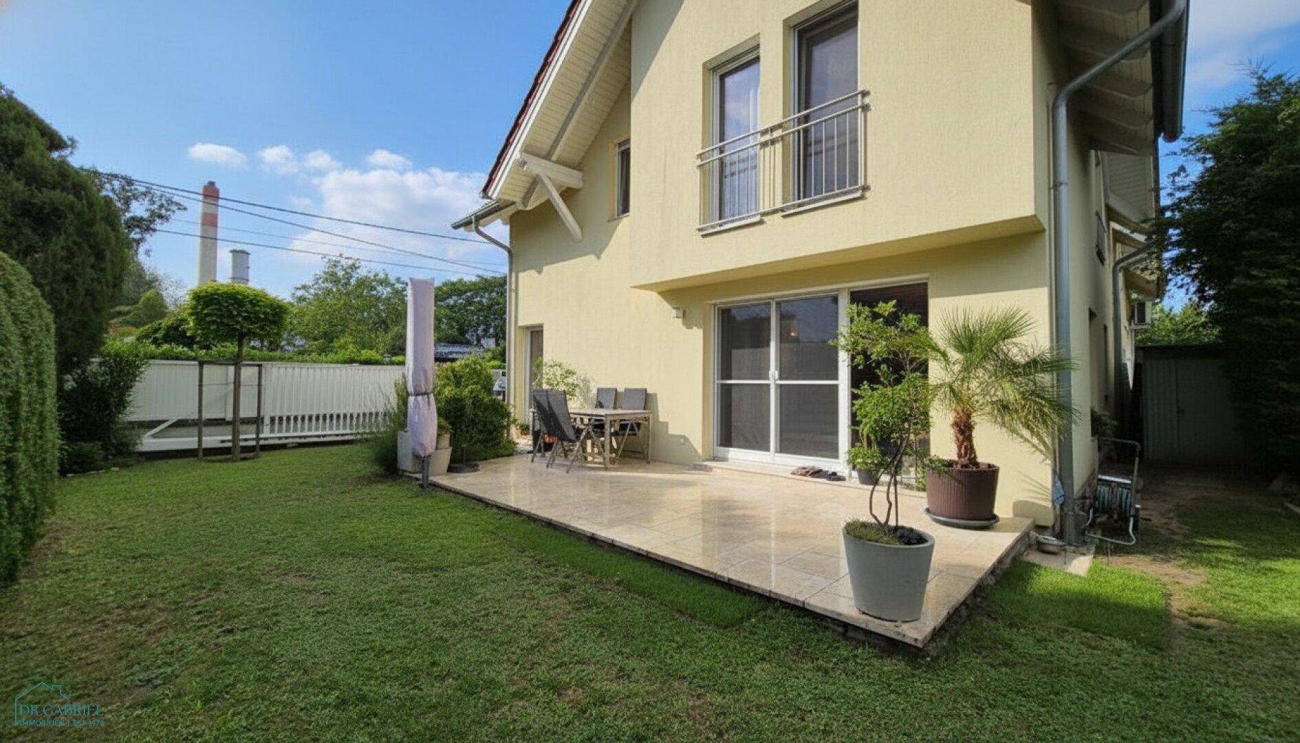 Hausfassade mit angrenzender Terrasse und weitläufigem Garten, umgeben von viel Grün.