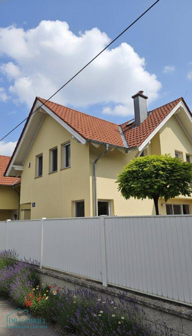 Moderne gelbe Hausfassade mit rotem Ziegeldach und einem gepflegten Vorgarten unter blauem Himmel.