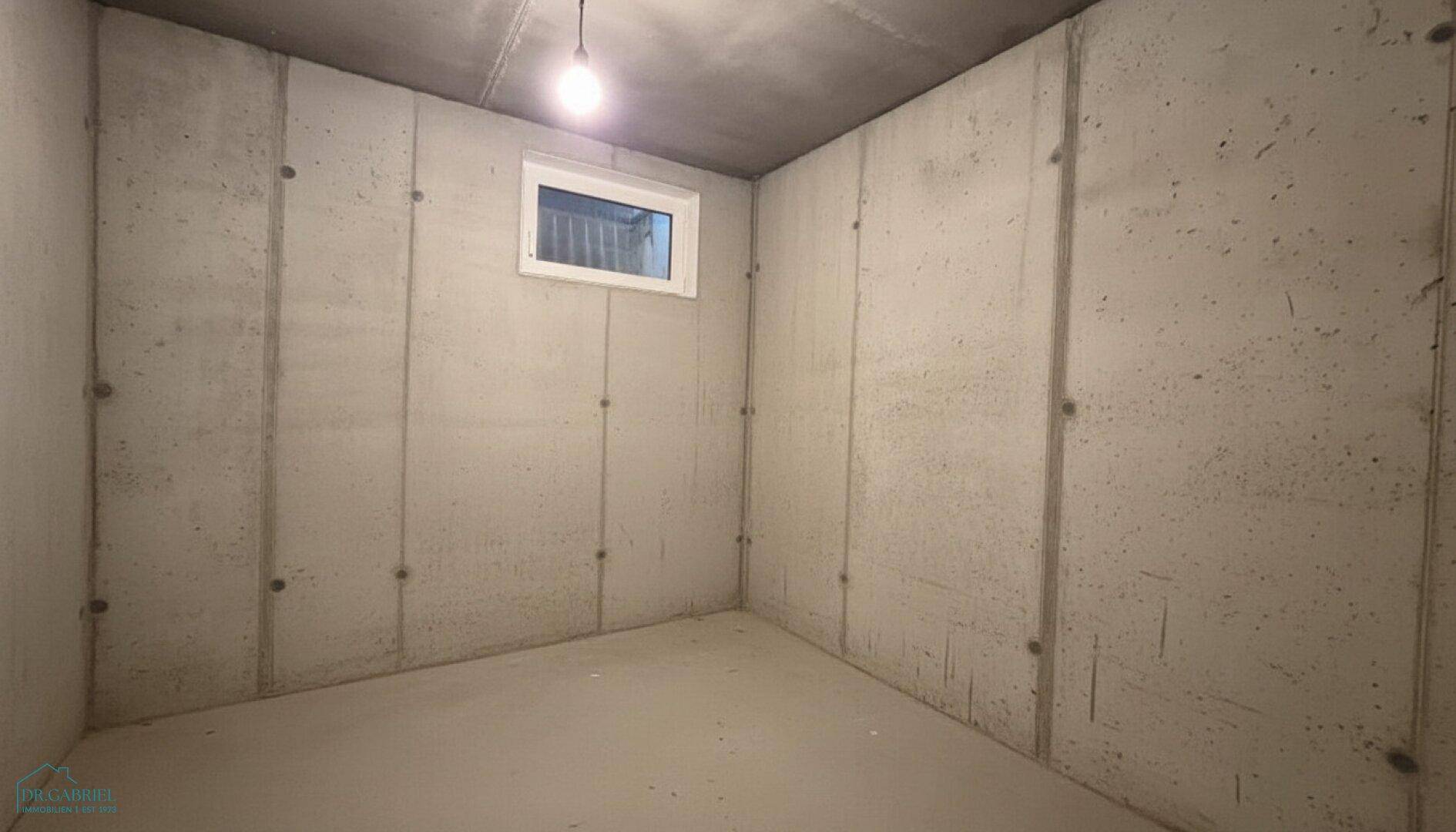 Unmöblierter Kellerraum mit rohen Betonwänden und einem kleinen, hochgelegenen Fenster für Belüftung.